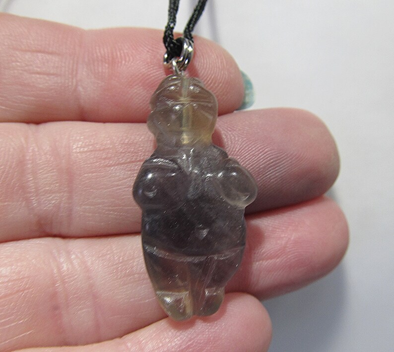 May include: A purple and clear crystal pendant carved in the shape of a human figure. The pendant is hanging from a black cord.