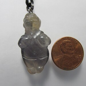 May include: A gray, translucent, carved stone pendant in the shape of a standing human figure. The pendant is about the size of a US penny.