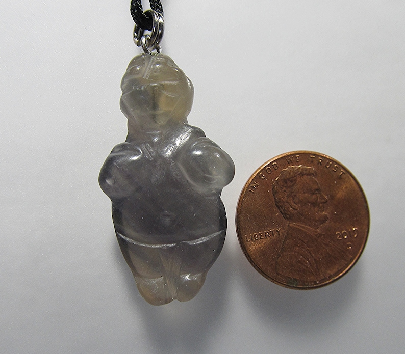 May include: A gray, translucent, carved stone pendant in the shape of a standing human figure. The pendant is about the size of a US penny.