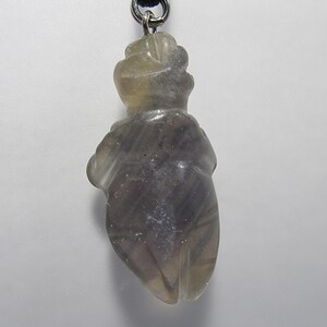 May include: A gray and white carved fluorite pendant in the shape of a human figure. The pendant is attached to a black cord.