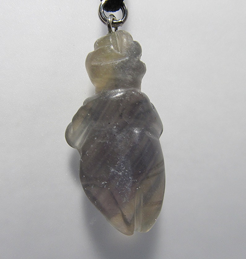 May include: A gray and white carved fluorite pendant in the shape of a human figure. The pendant is attached to a black cord.