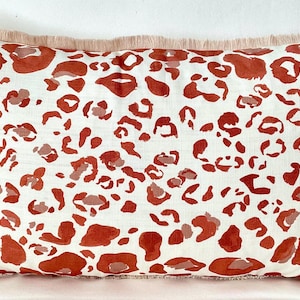 May include: Rectangular decorative pillow with a white background and a pattern of rust-colored leopard spots. The pillow has a light pink fringe trim around the edges. The pillow is designed for home decor.
