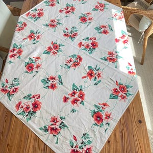 May include: A white tablecloth with a repeating floral pattern of red and pink flowers with green leaves and vines. The tablecloth is laid out on a wooden table, with the corners of the cloth angled towards the viewer.