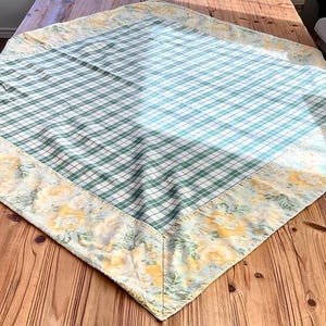 May include: A diamond-shaped tablecloth with a green and white plaid center. The border features a floral pattern with yellow roses on a light blue background. The tablecloth is placed on a wooden table.