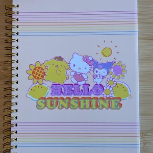 May include: A spiral-bound notebook with a light pink cover featuring Hello Kitty and friends, the text "Hello Sunshine," and rainbow stripes. Includes character-shaped page markers at the top. The notebook has gold-colored spiral binding.