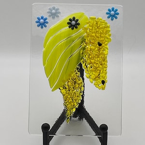 Puede incluir: Una obra de arte en vidrio fusionado con una hoja amarilla y racimos de cuentas de vidrio amarillas, acentuada con flores azules y negras. La obra se exhibe en un caballete de metal negro, mostrando un diseño único y artesanal.