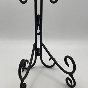 K&ouml;nnte beinhalten: Ein schwarzer Metall-Ausstellungsst&auml;nder mit einem dekorativen, gerollten Design. Der St&auml;nder hat einen dreibeinigen Sockel und eine Scharnierr&uuml;ckseite mit geschwungenen Akzenten an der Oberseite. Er dient zum Halten und Pr&auml;sentieren von Gegenst&auml;nden.