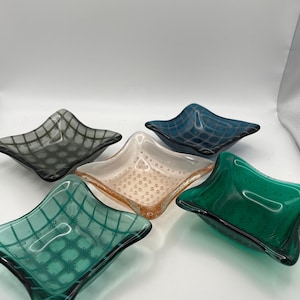 May include: Five colorful glass dishes in shades of green, blue, gray, and peach. Each dish has a square shape with curved edges and a grid pattern. The glass has a bubbly texture.