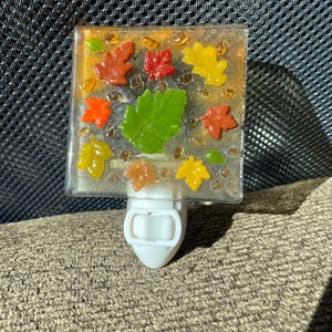 Peut inclure: Veilleuse décorative avec un panneau de verre carré orné de feuilles d'automne colorées. Les feuilles sont dans des tons de rouge, orange, jaune et vert, sur un fond clair. La veilleuse a une base blanche.