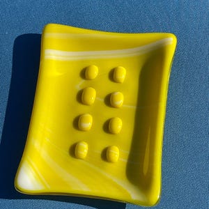 May include: A bright yellow glass soap dish with a rectangular shape and rounded edges. The dish features eight raised, oval-shaped bumps and white swirls. The dish is approximately 5 inches long and 4 inches wide.