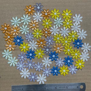 May include: A collection of colorful daisy-shaped embellishments in various colors, including orange, yellow, blue, white, and lavender. A metal ruler with inch measurements is placed near the flowers, showing the size of the embellishments.