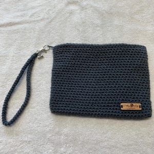 May include: A dark gray crocheted wristlet bag with a matching wrist strap and silver-tone clasp. The bag has a rectangular shape and a small wooden tag with text. The wrist strap is attached to the bag with a silver-tone clasp.