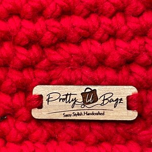 May include: Close-up of a vibrant red, chunky knit fabric. A small, rectangular wooden tag is attached, engraved with the words "Pretty U Bagz" and "Sassy. Stylish. Handcrafted."