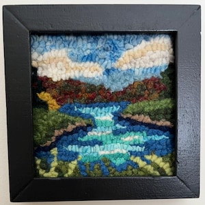 May include: A framed, textured artwork depicting a landscape scene. The piece features a blue sky with fluffy white clouds, a waterfall, and surrounding greenery. The artwork is encased in a black square frame.