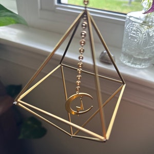 May include: A gold geometric pyramid with a crescent moon charm hanging from the top. The pyramid is made of thin metal rods and has a square base.