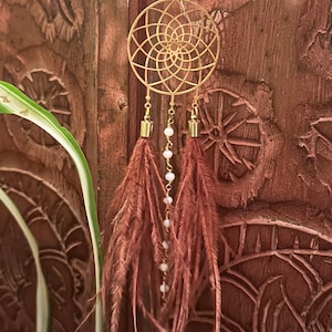 May include: A gold dreamcatcher with a circular design and brown feathers hanging from the bottom. The dreamcatcher is hanging from a chain with white beads.