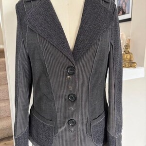 May include: A gray blazer with a textured collar and lapels, featuring a vertical striped pattern on the front panels. The blazer has four dark buttons and two front pockets. The garment is displayed on a mannequin.