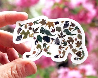 May include: A white sticker with a bear silhouette made of various leaves in shades of green, brown, and yellow. The sticker is held in a hand against a blurry background of pink flowers.