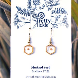 May include: Gold earrings with a small mustard seed encased in a clear hexagon. The earrings are on a white card with the text "Mustard Seed" and "Matthew 17:20" and the website address "www.theprettypickle.com".