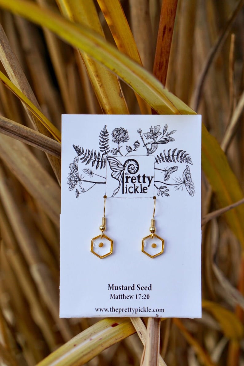 May include: Gold earrings with a small mustard seed encased in a clear resin hexagon. The earrings are on a white card with the text "Mustard Seed Matthew 17:20" and "www.theprettypickle.com".