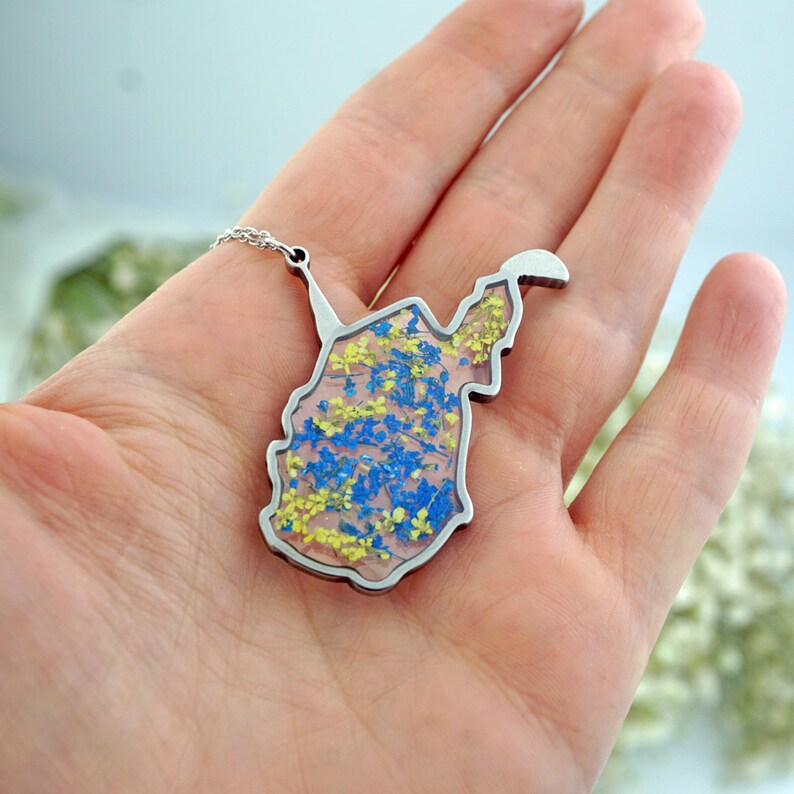 以下が含まれることがあります： 青い花と黄色い花が押し花された、ウェストバージニア州の輪郭のペンダントが付いたシルバーネックレス