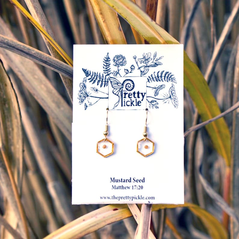 May include: Gold earrings with a small mustard seed encased in a clear hexagon. The earrings are on a white card with the text "Mustard Seed" and "Matthew 17:20" and the website address "www.theprettypickle.com".