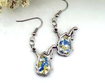 May include: A pair of silver earrings shaped like the state of West Virginia, with blue and yellow pressed flowers encased in clear resin.