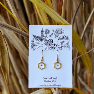 May include: Gold earrings with a small mustard seed encased in clear resin. The earrings are on a white card with the text "Mustard Seed" and "Matthew 17:20" and the website address "www.theprettypickle.com".