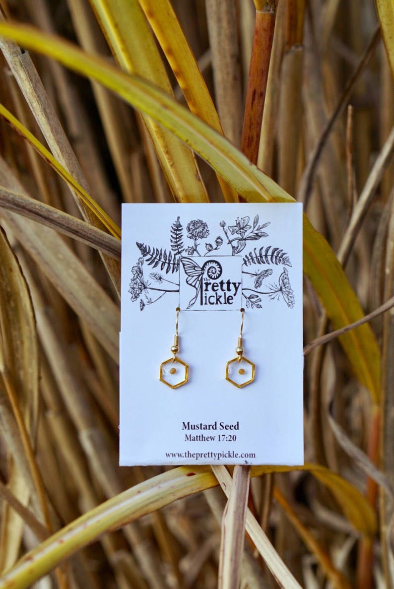 May include: Gold earrings with a small mustard seed encased in clear resin. The earrings are on a white card with the text "Mustard Seed" and "Matthew 17:20" and the website address "www.theprettypickle.com".
