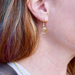 May include: Gold hexagonal earrings with a clear resin filled with a small, golden seed.