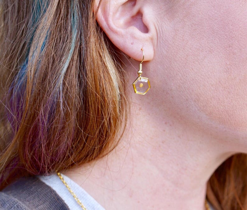 May include: Gold hexagonal earrings with a clear resin filled with a small, golden seed.