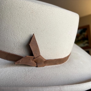May include: A cream-colored hat with a brown leather band and decorative knot. The hat has a wide brim and a structured crown. The leather band is secured with a decorative knot on the front of the hat. The hat is made of a soft, felt-like material.