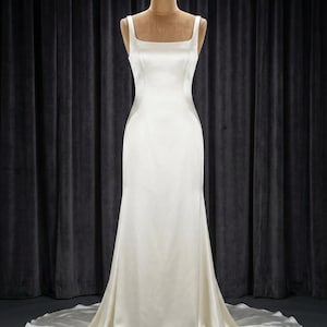 Può includere: Un abito lungo in raso color bianco sporco con scollo quadrato e spalline larghe. Il vestito presenta un corpetto aderente che si trasforma in una gonna fluida con un leggero strascico, esposto su un manichino per mostrare la silhouette.