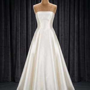 May include: A floor-length, strapless gown in a solid off-white colour. The dress features a fitted bodice that transitions into a full, A-line skirt with a smooth, satin-like finish. The garment is displayed on a mannequin with a neutral tone.