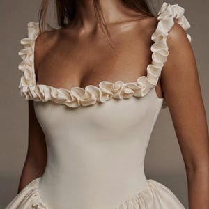 Op de afbeelding: Ivoor kleurige formele jurk met een getailleerd lijfje en een volle rok. De jurk heeft ruches langs de halslijn en bandjes, en decoratieve bloemaccenten op de rok. De stof lijkt een gladde satijn te zijn.