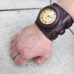 Puede incluir: Un reloj de pulsera con brazalete de cuero marrón con esfera crema y números negros. El reloj tiene una pequeña subesfera y se asegura con una hebilla. Un accesorio único.