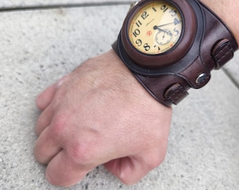 May include: A brown leather cuff watch with a cream face and black numerals. The watch has a small subdial and is secured with a buckle. The watch is a unique accessory.