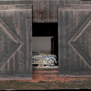 Può includere: Un'auto d'epoca parcheggiata all'interno di un fienile di legno con porte doppie. L'auto è ricoperta di polvere e detriti, e il fienile è buio e polveroso.