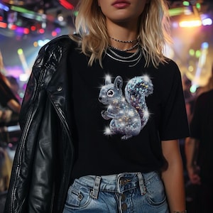 May include: A black t-shirt with a silver sparkly squirrel graphic. The squirrel is sitting on its haunches with its tail curled up behind it. The squirrel is surrounded by small, sparkly dots.