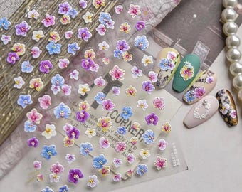 May include: Clear sheet of nail art stickers featuring small, 3D orchid flowers in shades of pink, purple, blue, and yellow. The stickers are arranged on a transparent backing, with some applied to painted nails. A string of pearls is visible.
