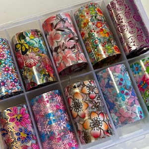 May include: A clear plastic organizer filled with ten rolls of nail art foil. Each roll features a different floral design in various colors, including pink, blue, orange, and purple. The foils are arranged in individual compartments.