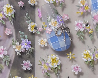 May include: A sheet of nail art stickers featuring a variety of floral designs. The stickers include white, yellow, pink, and purple flowers with green leaves, along with a butterfly and a nail with a blue and white checkered pattern.