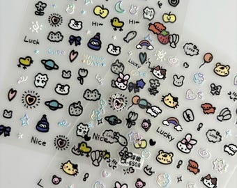 May include: Two transparent sheets of adhesive nail art stickers featuring a variety of small, colorful, and iridescent icons including cats, bears, strawberries, rainbows, planets, moons, stars, and text like Luck, Cute, Happy Day, and Nice.