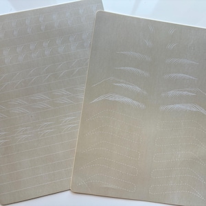 May include: Two beige practice sheets for eyebrow microblading. One sheet features various stroke patterns, while the other displays different eyebrow shapes. White dotted lines and feather-like strokes are visible on the sheets.