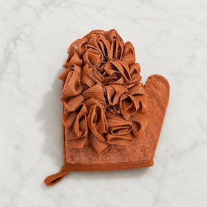Puede incluir: Un guante de baño de color terracota con una parte superior decorativa con volantes. El guante tiene un lazo para colgar y está hecho de un material texturizado. El guante está diseñado para usar en el baño o la ducha.