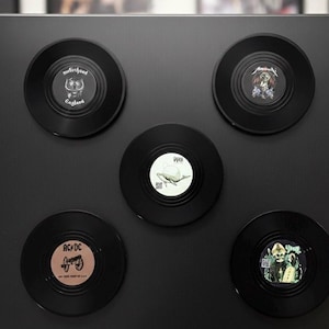 Può includere: Un set di sottobicchieri in vinile nero con copertine di album di band rock come Motörhead, Metallica e AC/DC. I sottobicchieri sono disposti su una superficie nera.