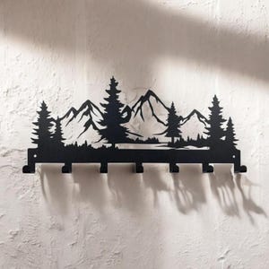 May include: Black metal wall-mounted coat rack with a silhouette of a mountain range and trees. The rack has seven hooks.