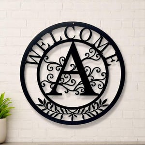 May include: Black metal welcome sign with the letter "A" in the center, surrounded by decorative scrollwork and the word "WELCOME". A circular design with a leaf border at the bottom. The sign is mounted on a white brick wall.