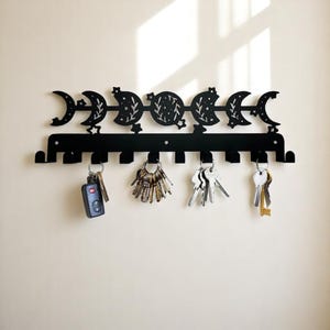 May include: Black metal key holder with a celestial moon phase design. The key holder has hooks for keys and is mounted on a wall. Several sets of keys hang from the hooks, including a car key fob.