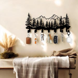 May include: Black metal key holder shaped like a mountain range with pine trees. The holder has six hooks for hanging keys, and there are keys and other items hanging from the hooks.